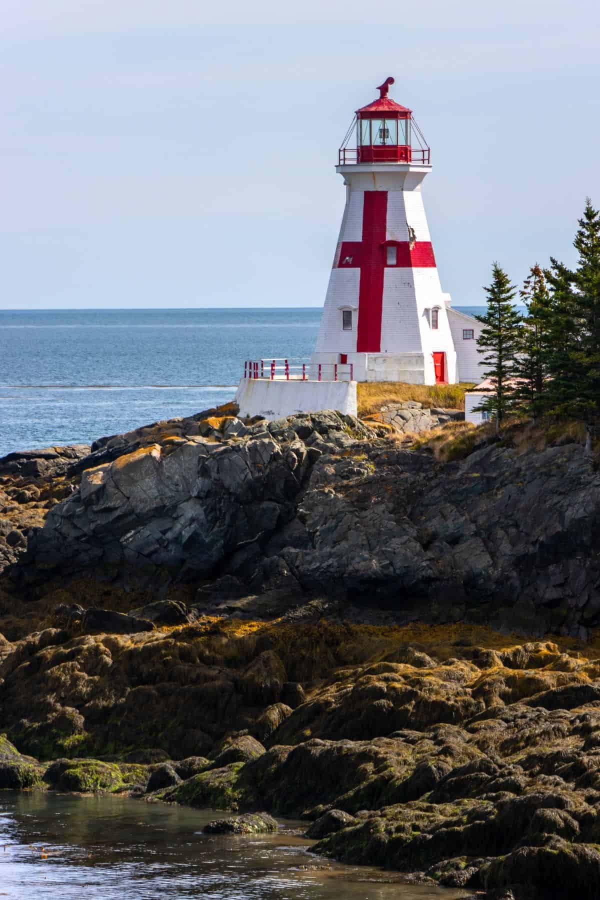 Red and white striped lighthouse on rocky coastal outcrop with ocean and evergreen trees in background