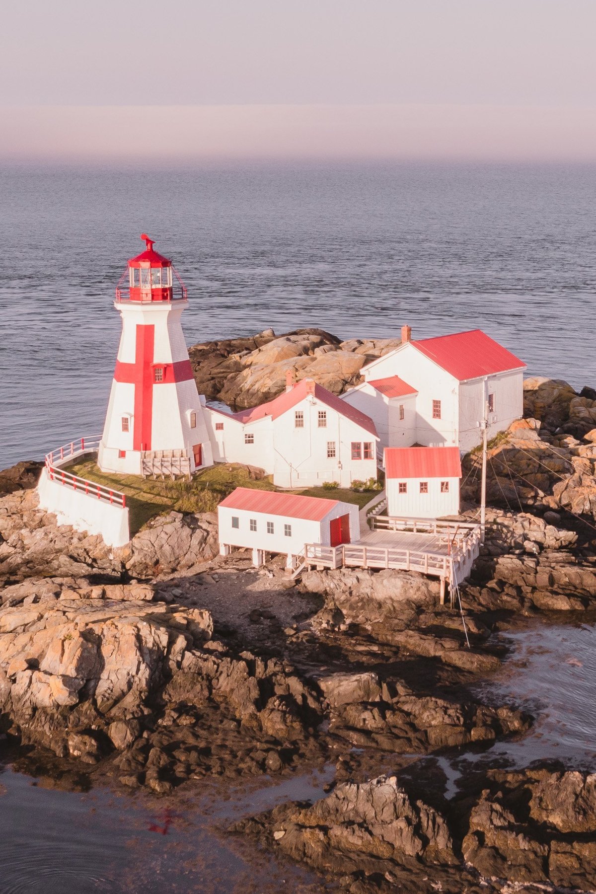 Red and white lighthouse with keepers buildings on rocky outcrop surrounded by ocean at dusk